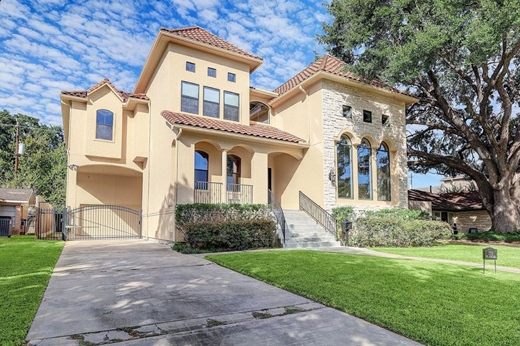 A multi-story, cream-colored home in the Braeswood neighborhood in Houston, Texas. There are multiple windows and a lush green yard. 