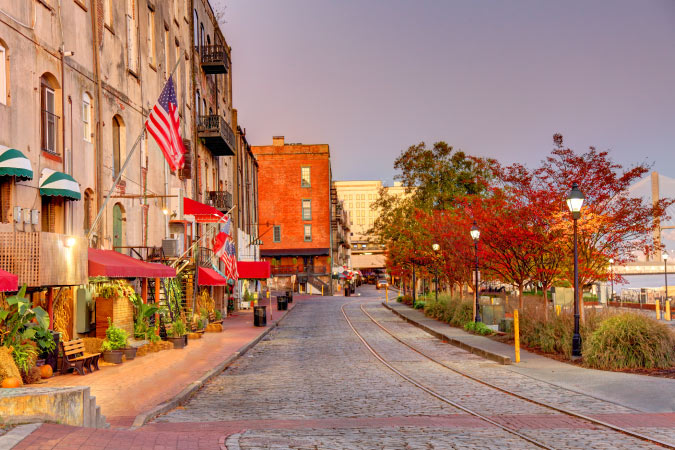 View of Savannah’s historic River Street at dusk, featuring historic buildings, a cobblestone road, and a bridge in the distance.