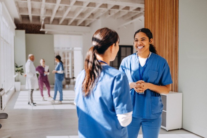 Two nurses are smiling while talking in a hallway while three other healthcare workers are doing the same in the background.