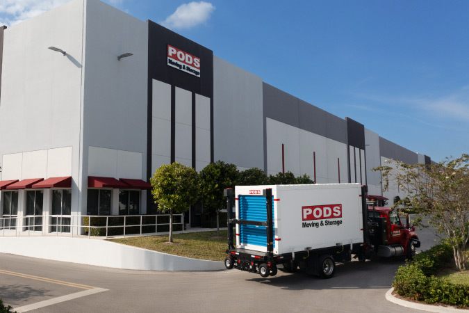 A PODS truck is delivering a portable storage container to a PODS storage facility, pulling into the parking lot before the container is unloaded.