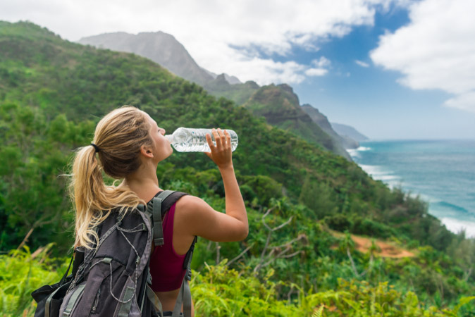 A woman is drinking from a bottle of water during a hike in Hawaii, with a stunning view of lush mountainside and the blue Pacific ocean ahead of her.