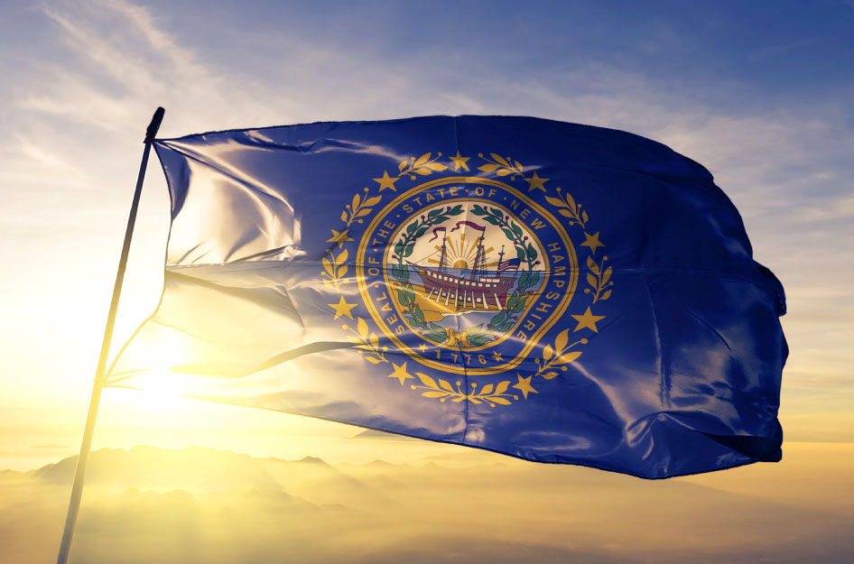 The New Hampshire state flag is flying on a pole against a picturesque backdrop of mountains and a setting sun. The flag consists of a blue background with the state's seal at its center. The seal showcases the frigate Raleigh surrounded by laurel leaves and nine stars.