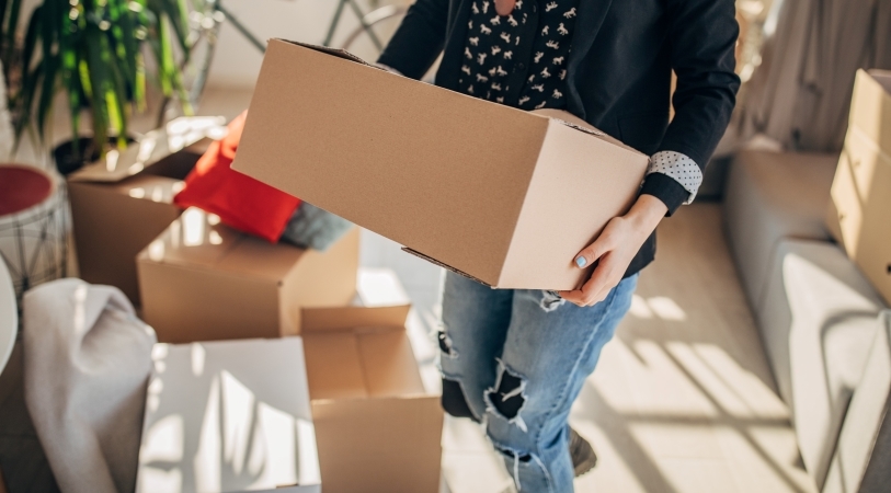 Close-up view of a stylish woman carrying a moving box out of her apartment. The room behind her has several other moving boxes, as well as some furniture and a houseplant. 