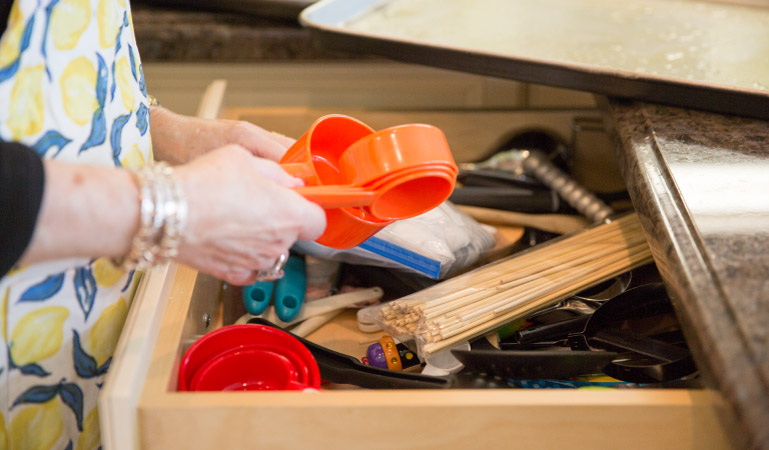 A woman organizes measuring cups by size, stacking them in each other to save space. The drawer she’s working out of contains wooden sticks, pliers, and other various items.