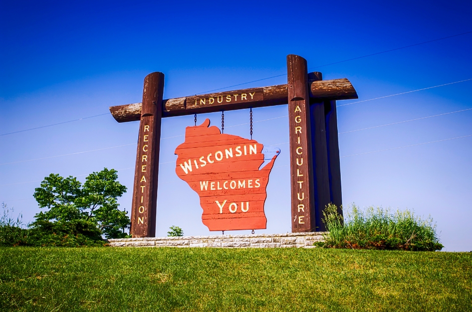 A large, red Wisconsin-shaped welcome sign reading “Wisconsin Welcomes You,” supported by wooden posts highlighting key aspects of Wisconsin life — “recreation,” “industry,” and “agriculture.”
