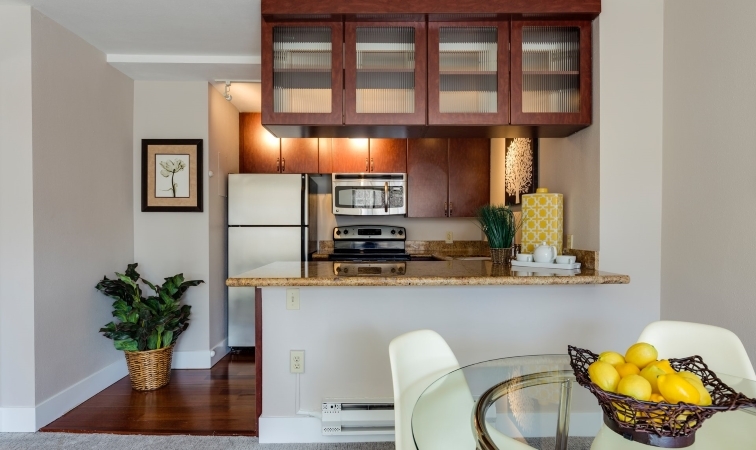 View of a small kitchen from the dining area of a small apartment. In addition to typical appliances, the kitchen features an island/bar with overhead storage. 