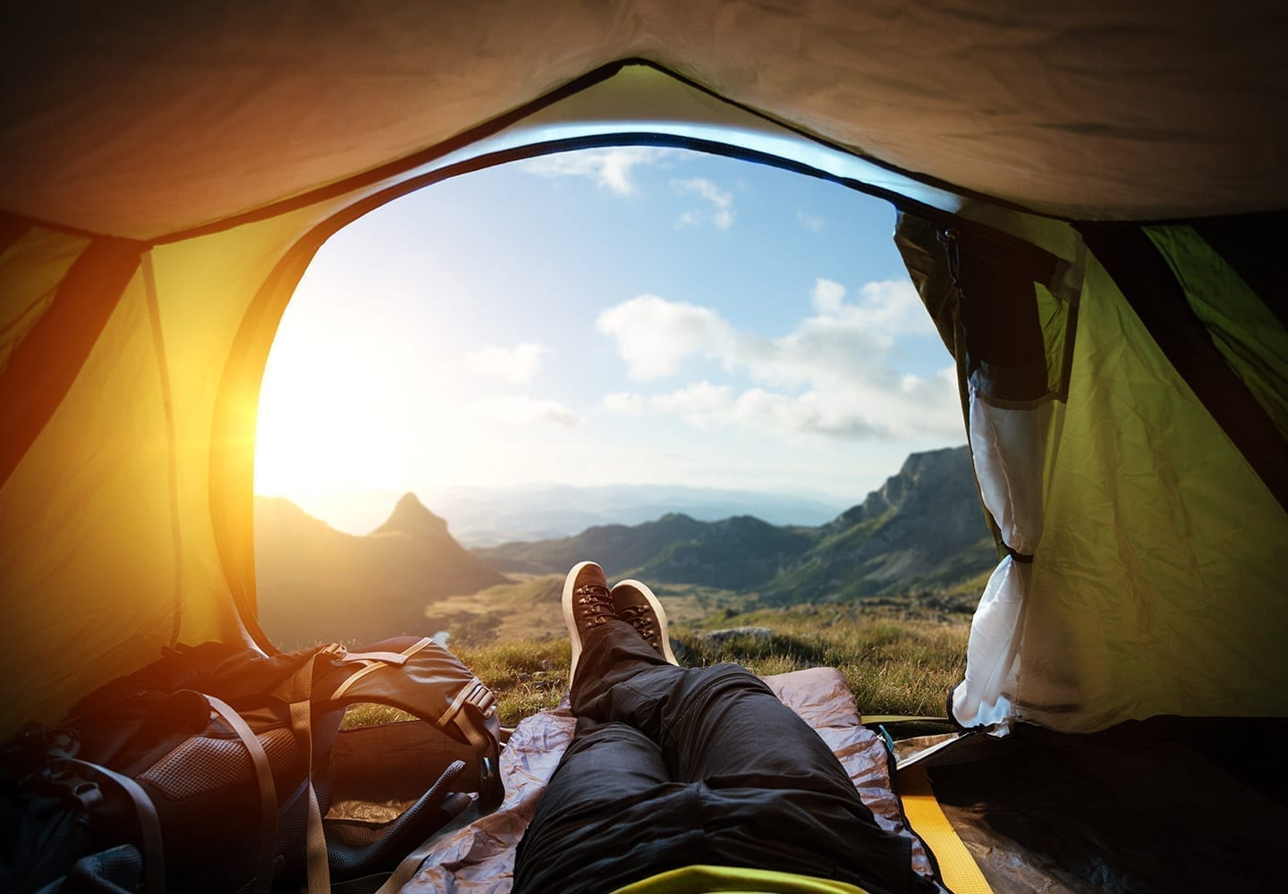 A camper wearing jeans and climbing shoes relaxes in his yellow tent overlooking the mountains at sunset