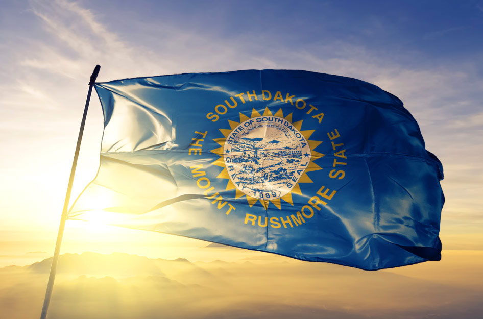 The South Dakota state flag, blowing in the wind at the top of a simple flag pole. Behind the flag, the sun sets behind a low range of mountains.