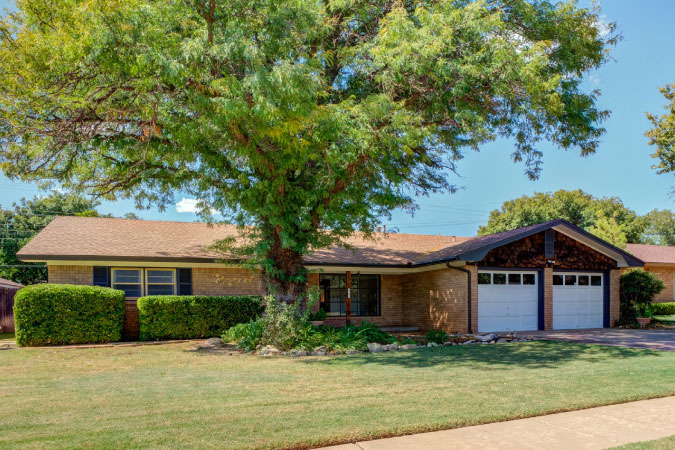 A two-bedroom, single-story home in Melonie Park — one of the best Lubbock neighborhoods — featuring a gabled garage with two garage doors and a large tree in the front yard.