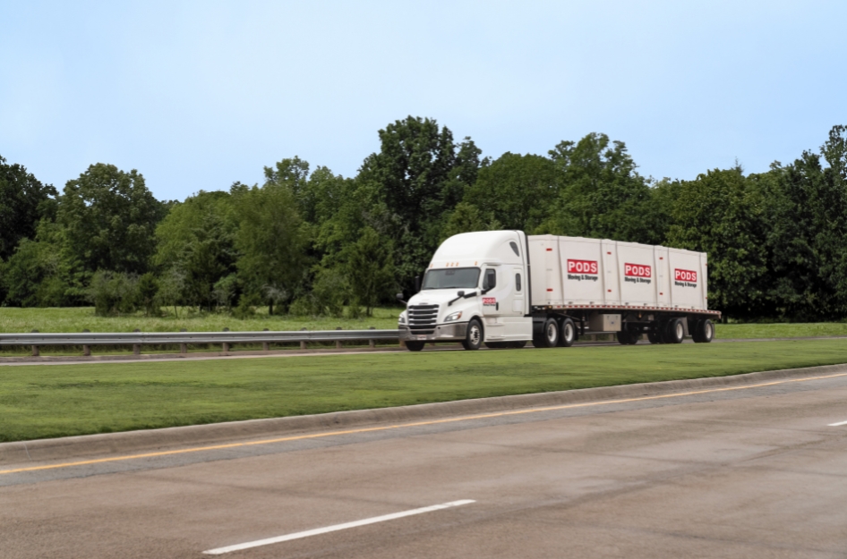 A PODS truck is transporting three portable moving and storage containers along a freeway, past lush forests and fields, as part of a long-distance move.