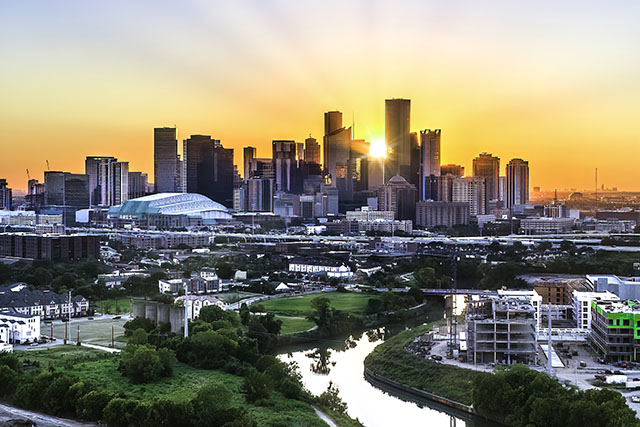 A panoramic view of the Houston, Texas, skyline. The sun peeks out from behind skyrise buildings. The city’s football stadium is visible in the background.