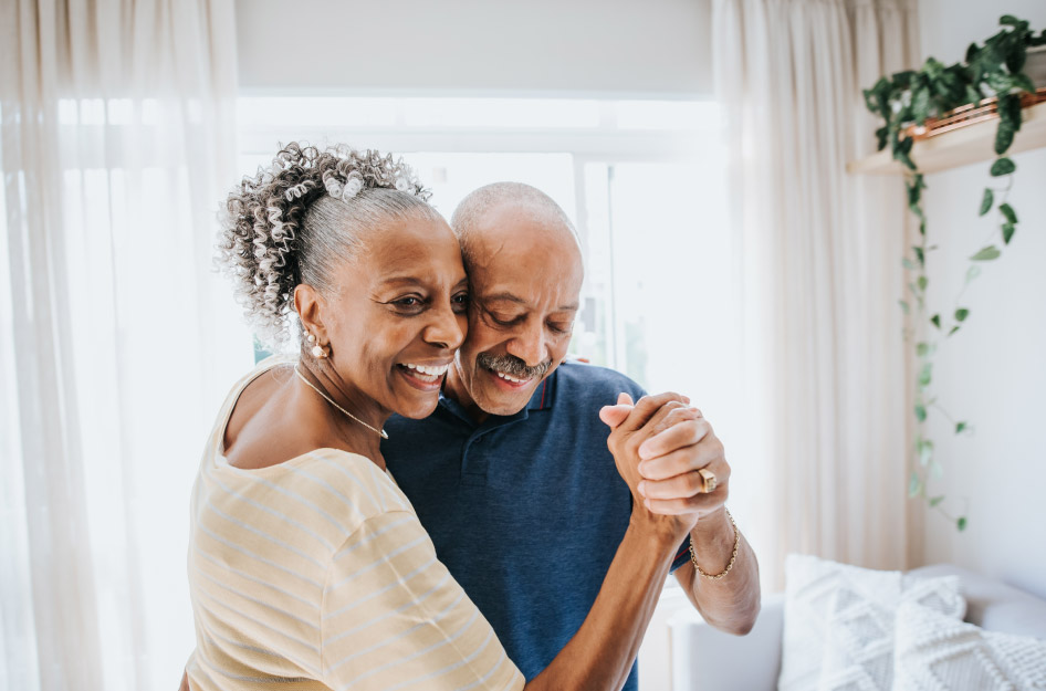 A senior couple is dancing in their living room as the sun shines in through the window behind them.
