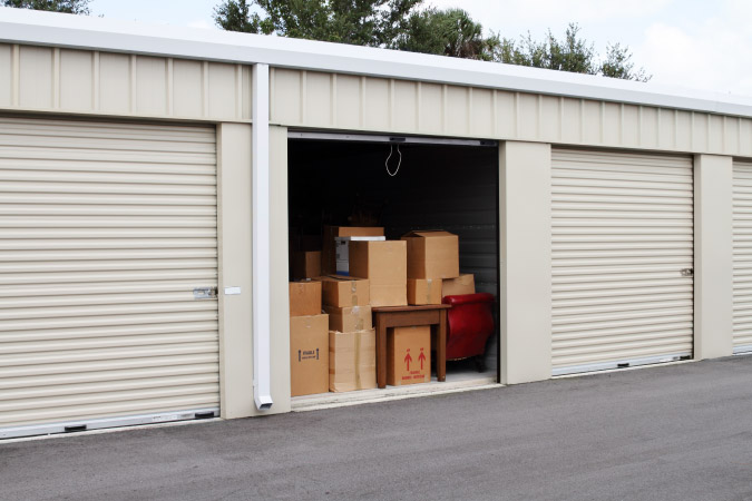 Exterior view of a row of self-storage units with the center one open, revealing a couple of pieces of furniture and a dozen cardboard boxes.