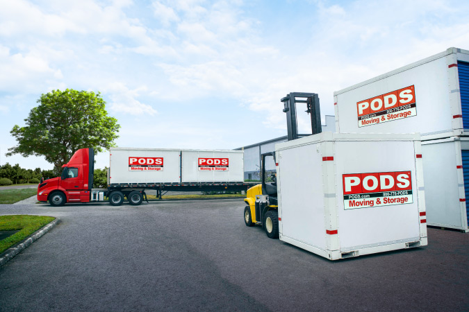 View of the side of a PODS storage center where a truck is about to depart with two PODS containers and a forklift is moving another container.