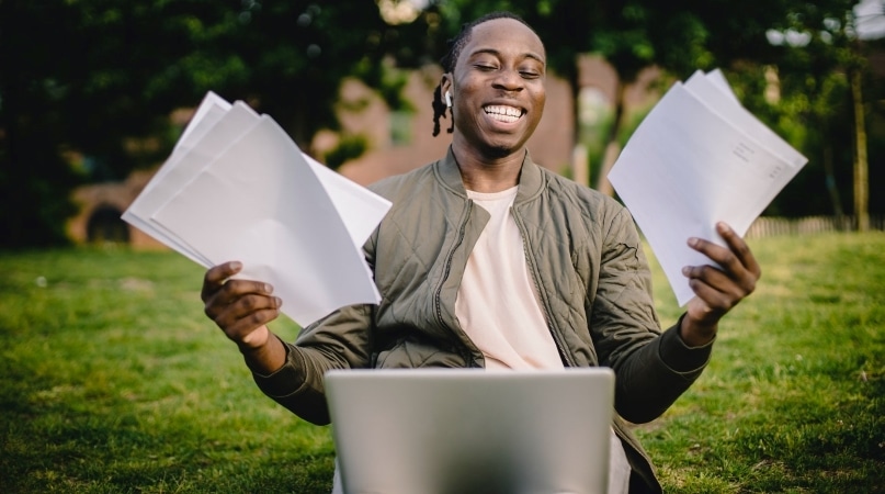 A man smiles while holding multiple moving estimates in his hands and researching moving companies on his laptop.
