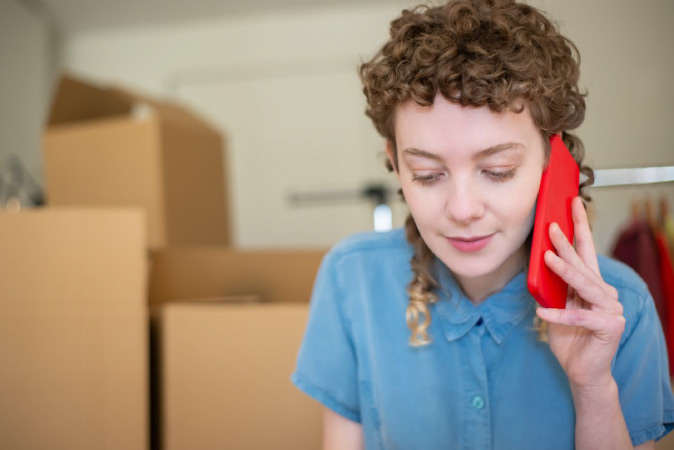 A woman is speaking on her cell phone with moving boxes stacked behind her.