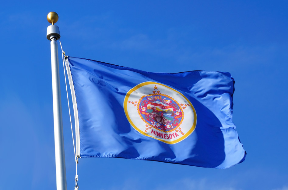 The Minnesota state flag is blowing in the wind at the top of a flagpole in front of a clear, blue sky. The flag is blue with a gold-lined circle in the middle holding the state seal.
