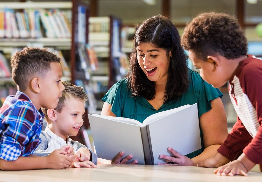 A librarian sits at a table and reads to three young boys . You can see the shelves of different books in the background. 