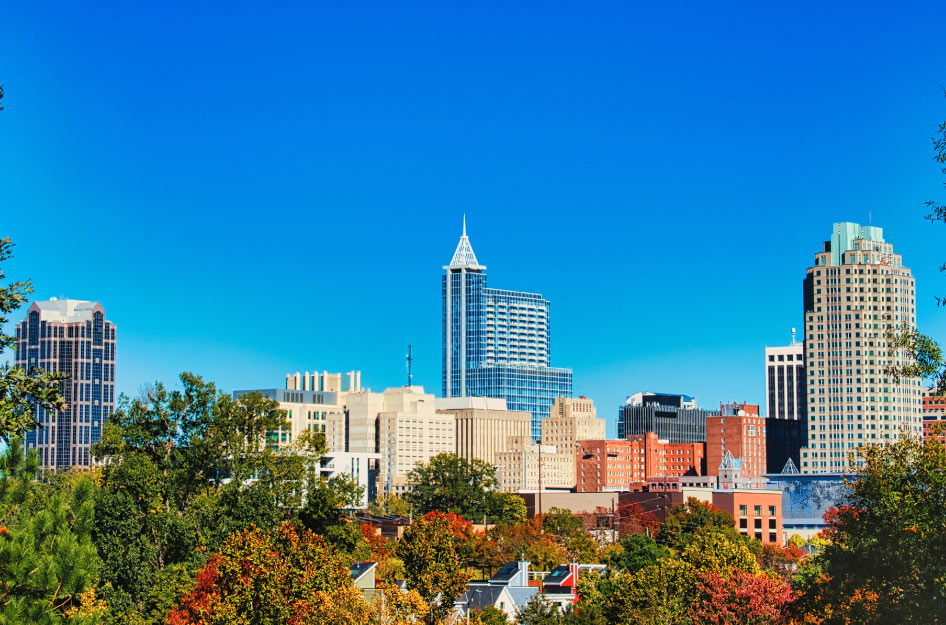 Downtown Raleigh skyscrapers, with fall-colored leafy trees in the foreground and a bright blue, cloudless sky in the background.