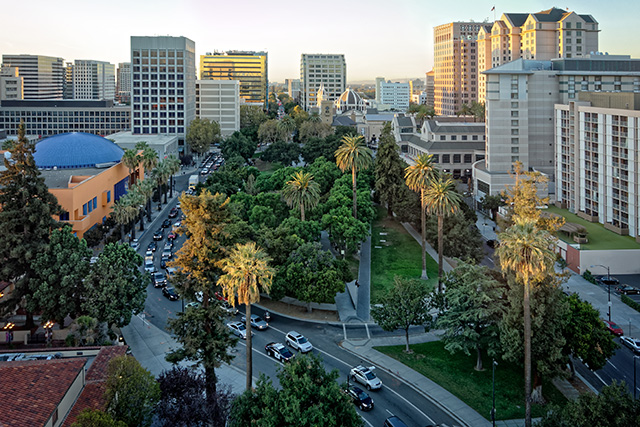 The Plaza de Cesar Chavez in San Jose, California, during the daytime. There are two lanes of full traffic and green palm trees surrounded by buildings.