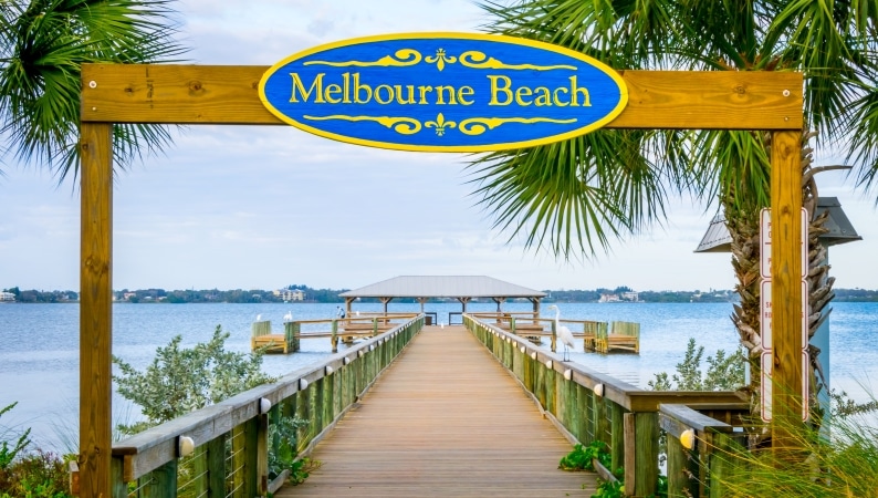 A long pier in Melbourne, Florida, reaches out into the Indian River. A blue and yellow sign is posted above the pier and reads, “Melbourne Beach.”