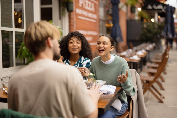 Three friends are laughing together while sharing an al fresco meal at a city restaurant.