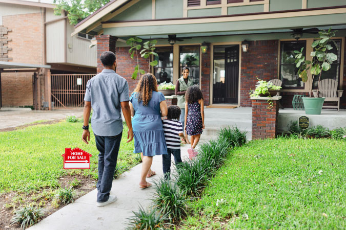 A family of three is up the entry path to a Texas bungalow with a “Home For Sale” sign in the front yard.