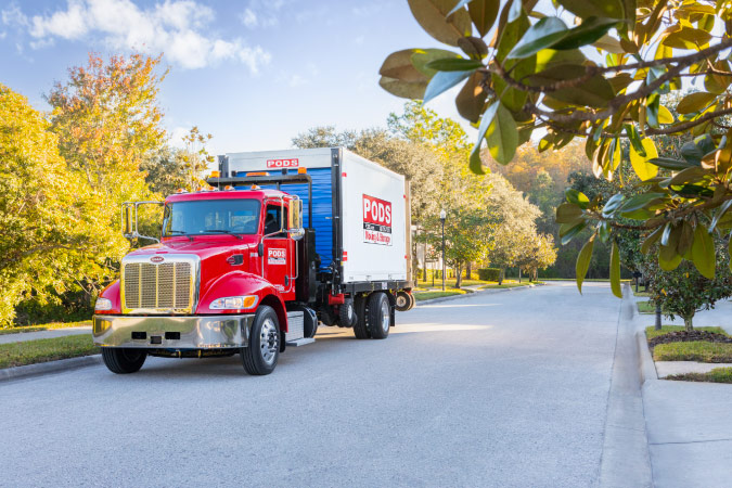 A PODS truck is transporting a PODS portable moving container through a neighborhood to a residential home.