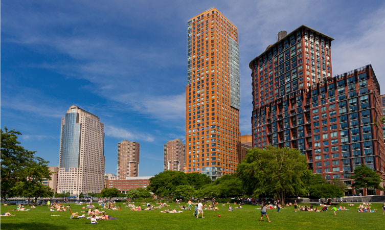 Dozens of locals are enjoying a sunny day outdoors in Battery Park City, New York, one of the safest neighborhoods in NYC, with tall residential buildings rising up in the background.