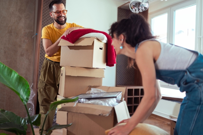 A smiling couple moving during hurricane season stands among stacked moving boxes inside their home as they prepare for an upcoming move.