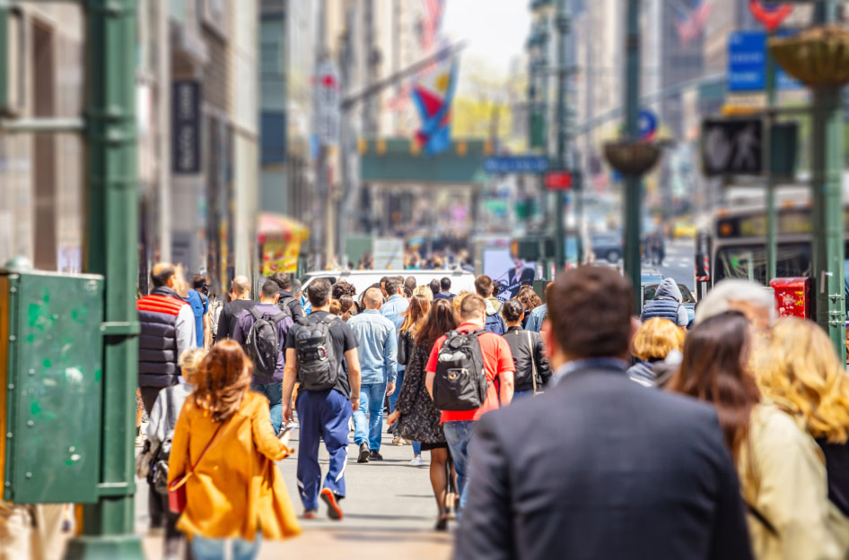 Street-level view of crowds of pedestrians crossing a street in New York City on a sunny day.