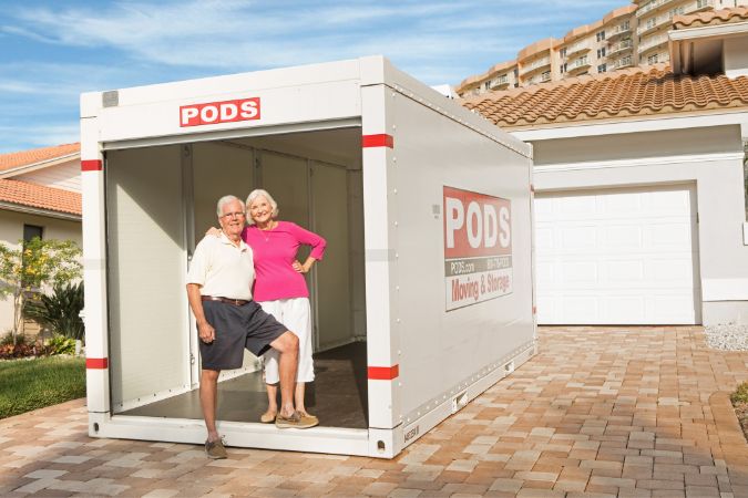 A senior couple is standing together in the front of an empty, open PODS storage container in their driveway.