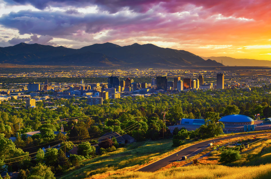 Distant view of Salt Lake City and the surrounding mountains at sunset. The sky is full of clouds varying in color from deep purple to bright yellow. Downtown buildings stand tall above the residential areas around them. In the foreground is a grass-covered hill and a thick forest of trees.