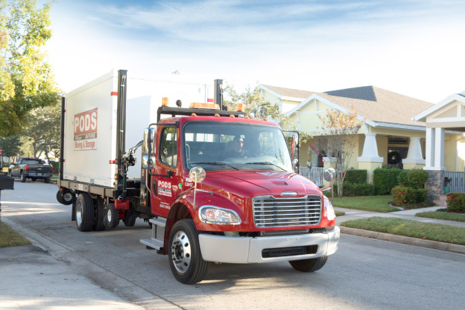 A PODS truck is transporting a portable storage container through a residential neighborhood to a customer’s home as part of PODS’ self-storage with delivery service.