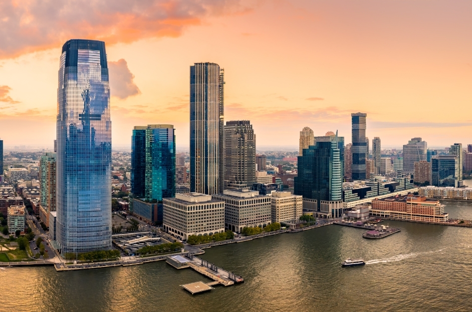 Sunset view of the Jersey City skyline along the Hudson River with high-rises and a ferry boat moving across the water, highlighting the vibrant waterfront and diverse Jersey City neighborhoods.