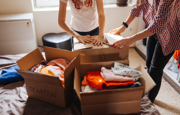 A mother and daughter are closing one of three cardboard boxes filled with clothing donations. They’re decluttering and minimizing their belongings to save on interstate moving costs