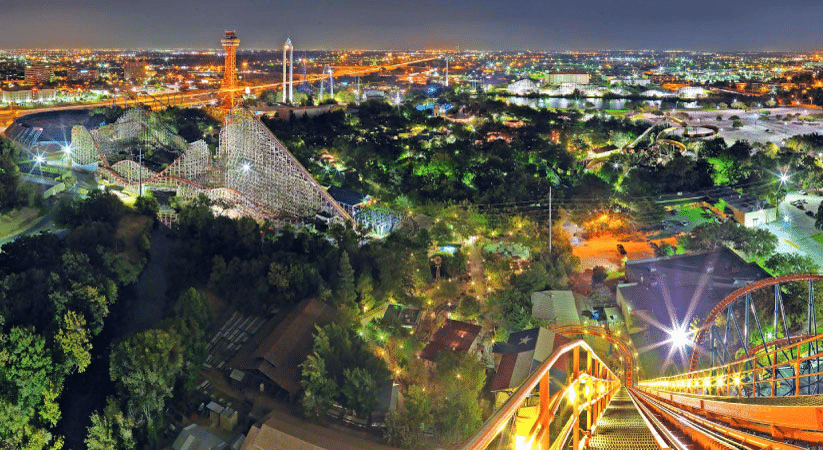 An evening view from the top of a roller coaster at Six Flags Over Texas, featuring a vista of the surrounding city of Arlington, illuminated by street lights, residential buildings, and cars. 