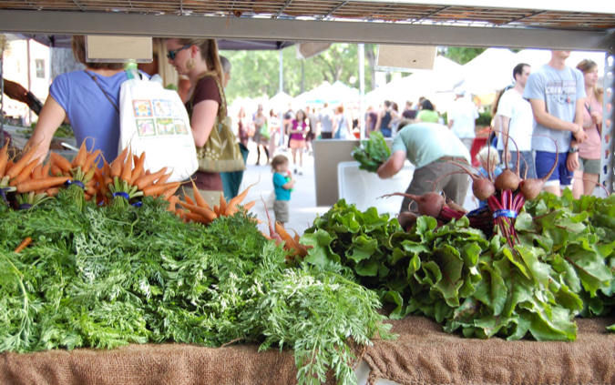 Bunches of carrots and beets fill a table at a farmer’s market stand in the foreground while people living in Boulder, CO, explore tents at a farmers’ market.