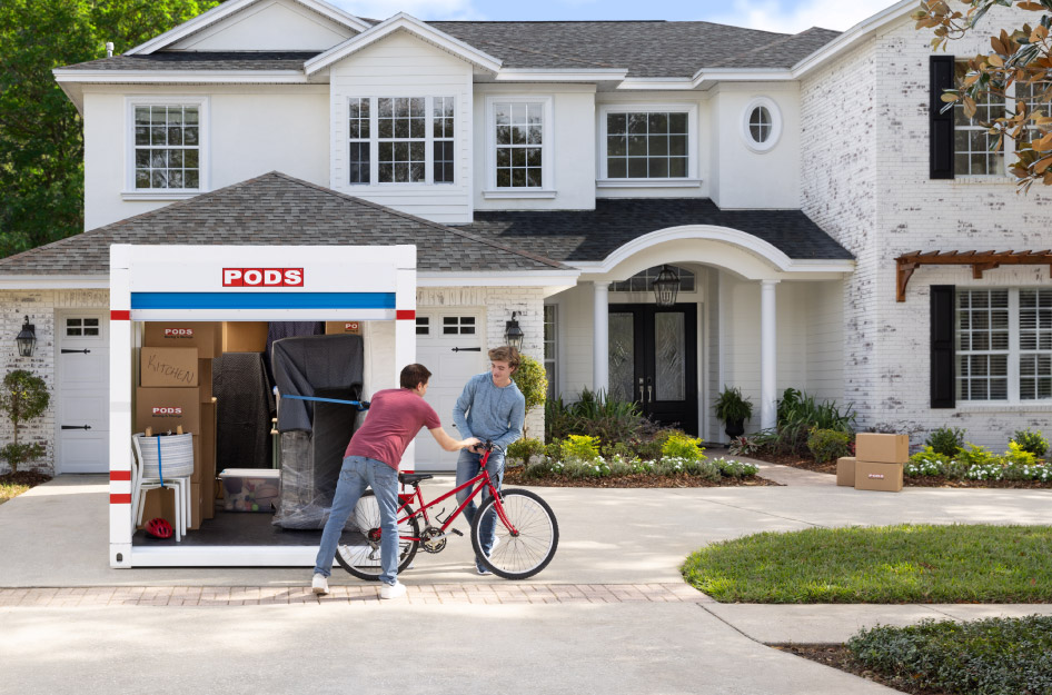 After renting a container for moving, a father is handing his son a red bicycle as they unload the container in the driveway of their new house.
