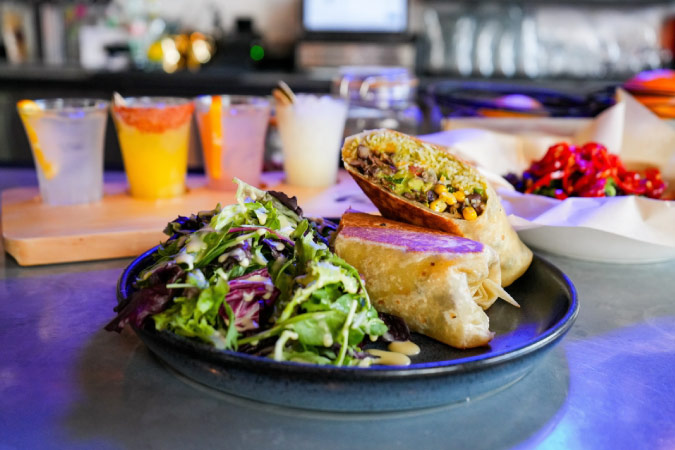 A close-up view of a burrito and side salad with a flight of special cocktails in the background at Chido Taco Lounge in Frisco, Texas. 