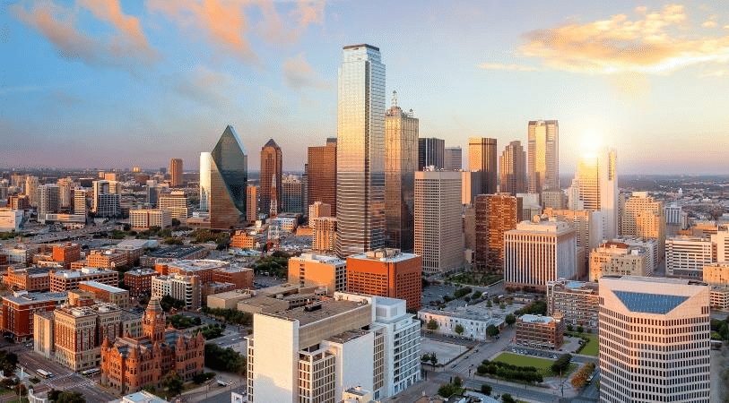 In an aerial view of Downtown Dallas during sunrise, the city’s glass skyscrapers reflect the sky in a spectrum of colors from soft peaches to light blues.