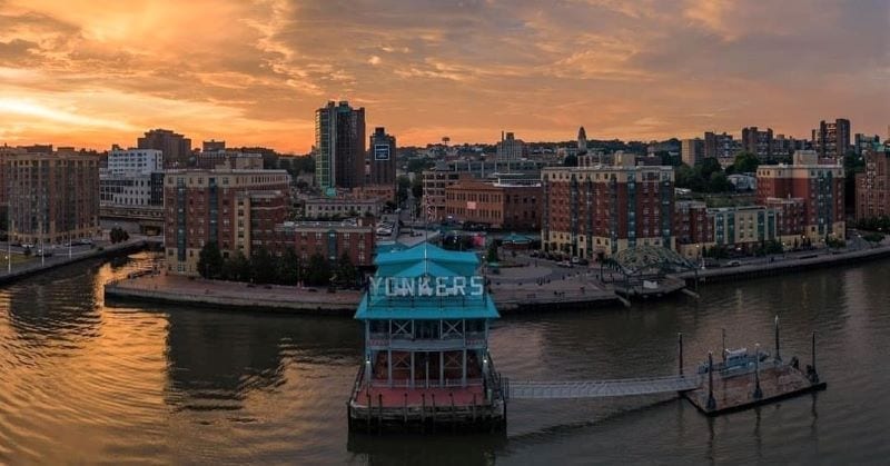 Aerial view of the Yonkers downtown waterfront at sunset. The water is mostly calm, as light orange hues in the sky are mirrored below.