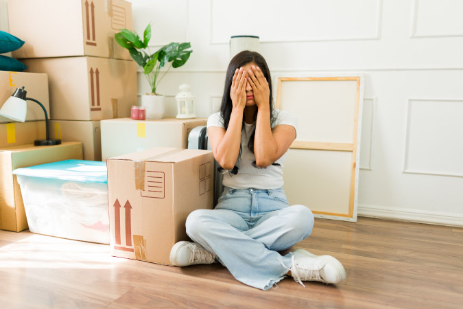 A stressed woman is sitting on the floor beside moving boxes and other household items, holding her hands to her face in exasperation.