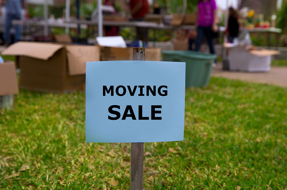 A simple paper sign with the words “MOVING SALE” printed in black letters is posted in the front yard of a residential home. In the background, tables and boxes are set up with items on display.