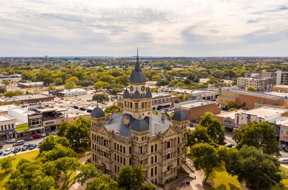 Aerial view of Denton, Texas — one of the cities near Dallas — featuring a city building with a lot of character, surrounded by mature trees and city shops.