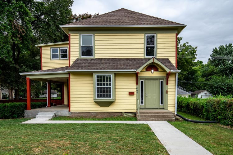 A cute two-story home in the small town of St. Marys, Kansas. Th house is painted a light yellow with green and red trim. The front door is green and the covered porch features matching red columns. 