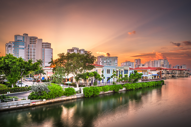 The skyline of Naples, Florida, at sunset. There are colorful shops on the water surrounded by different greenery and shrubs. The sun peeks out of the top of the image.
