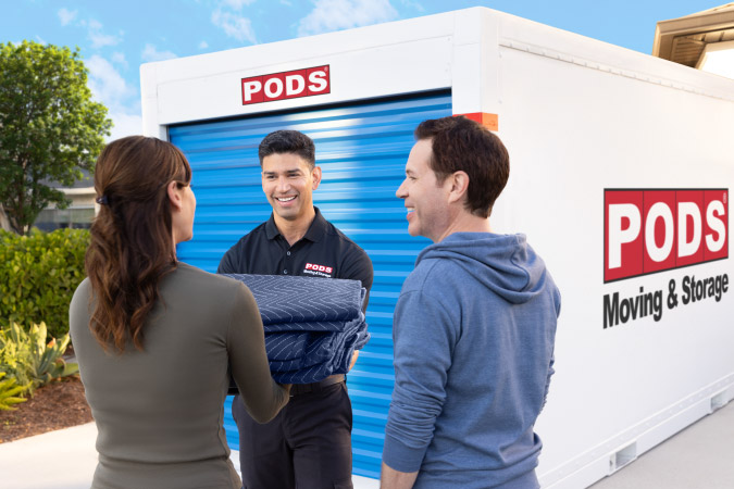 A PODS driver is handing moving blankets to a couple beside a PODS portable moving container in their driveway.