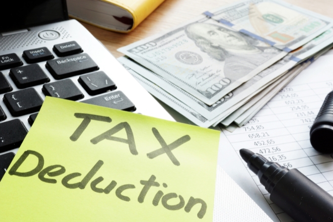 Close-up of the desk of someone who’s been calculating their moving expenses tax deduction, including financial documents, cash, a laptop, and a sticky note with the words “TAX Deduction” written on it.