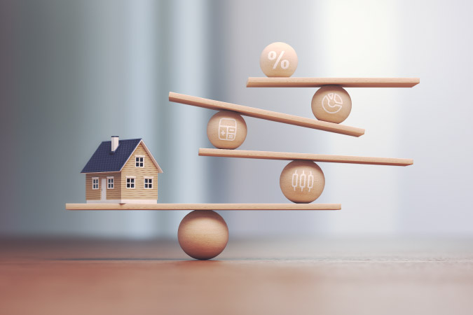 A small model of a house and several small wooden balls balanced delicately on wooden boards, symbolizing the delicate balance between interest rates and inflation.